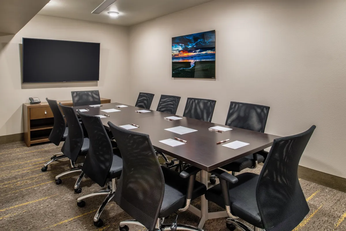 This image showcases a spacious, modern conference room with a large conference table surrounded by ergonomic office chairs. A wall-mounted television screen is visible, ideal for presentations or video calls, and framed artwork adds a touch of sophistication. The atmosphere is collaborative, reflecting a space well-suited for meetings, brainstorming sessions, or networking.