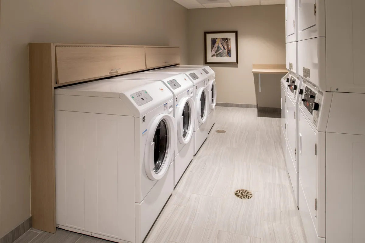 A clean and practical laundry area featuring multiple washing machines and dryers, counter space for folding clothes, and decorative artwork. The efficient layout offers both functionality and a sense of comfort for longer-stay guests.