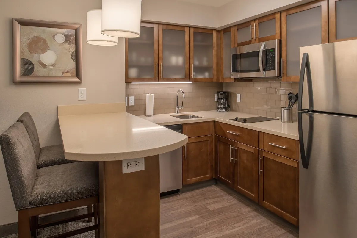 A warmly lit interior scene featuring a bar seating area adjacent to a modern kitchen with up-to-date appliances. Guests are seen preparing food and socializing, evoking a sense of togetherness. Artwork adorns the wall, and the open concept design encourages connection between the kitchen and dining spaces.