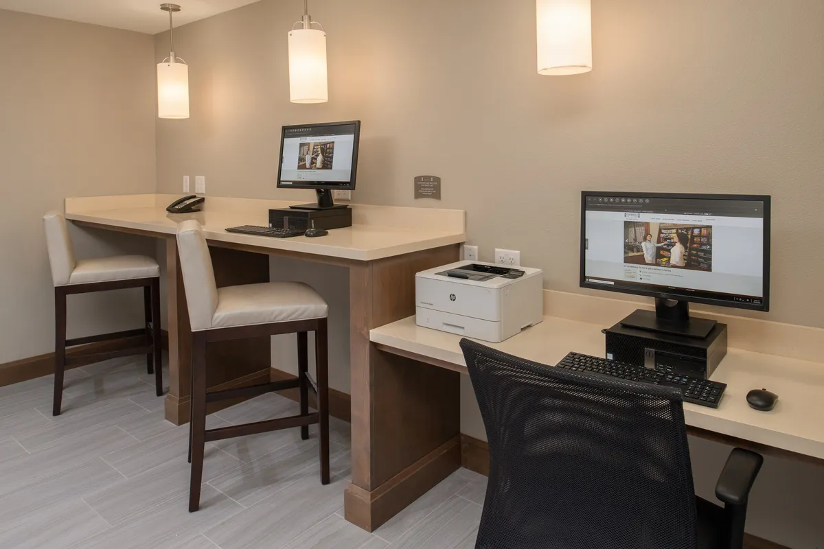 The image features individual computer workstations, a printer, and stylish lighting fixtures, creating an environment tailored for focused work. The setup enables guests to catch up on tasks, print documents, or conduct virtual meetings in a quiet, professional setting. The mood is one of concentration and efficiency.