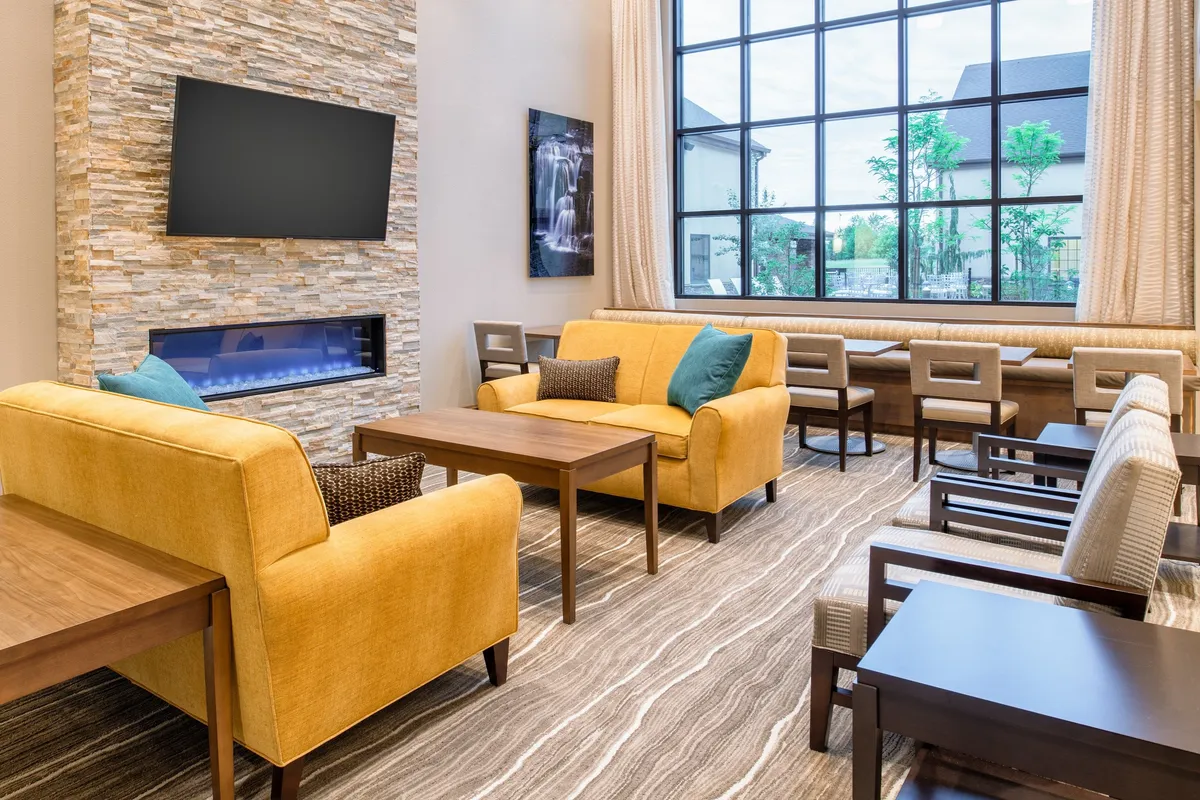A spacious living area with vibrant yellow sofas arranged around a stone fireplace, large window flooding the space with sunlight, and tasteful decorative artwork. The room offers both comfort for relaxation and a designated area for work or socializing.
