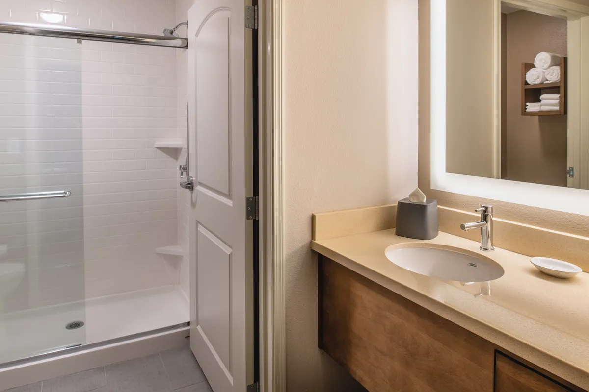 A contemporary bathroom featuring a glass-enclosed shower, sleek sink and vanity with a large mirror, and a shelf neatly stocked with fresh towels. Warm lighting and clean lines evoke a spa-like atmosphere.