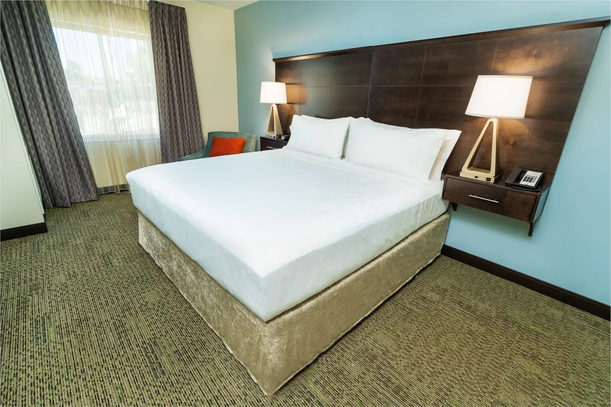 A peaceful, well-lit bedroom featuring a plush king-size bed positioned near a window, with a nightstand and lamp providing a cozy nook for reading or relaxing. The neutral décor and inviting linens create a restful and stylish retreat.