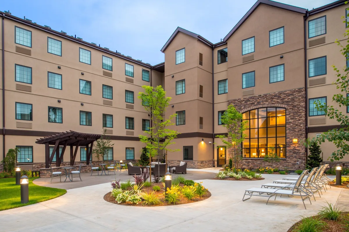 The image shows a welcoming courtyard seating area surrounded by landscaped gardens, with the large windows of the Staybridge Hillsboro North building as a backdrop. The setting captures a tranquil, inviting atmosphere—ideal for relaxing or enjoying family time outdoors, with sunset lighting adding warmth and comfort.