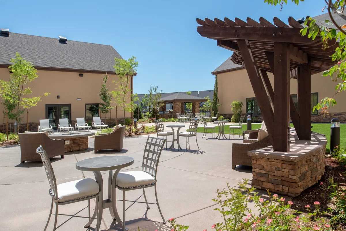 An inviting outdoor patio space with comfortable seating beneath a pergola, surrounded by landscaped greenery. The area is set up for social gatherings, with guests relaxing, enjoying meals, and taking in the cheerful ambiance—perfect for an evening BBQ or casual dining experience.